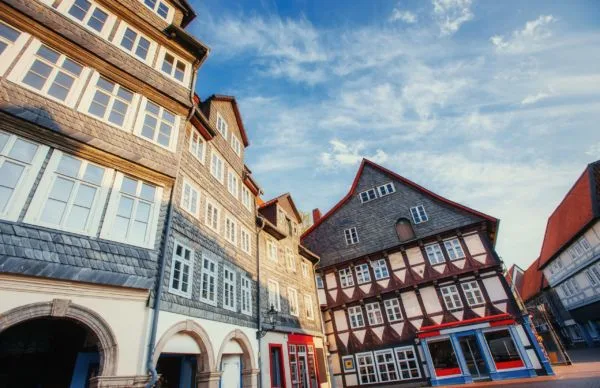 Eine Froschperspektive einer Reihe traditioneller deutscher Fachwerkhäuser unter einem strahlend blauen Himmel mit weißen Wolken. Die Gebäude haben unterschiedliche architektonische Stile, eines davon mit dunklen Schindeln und gewölbten Eingängen, ein anderes mit einem markanten Giebeldach und schwarz-weißem Fachwerk