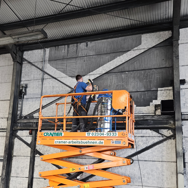 Ein PUR-Mitarbeiter in blauer Kleidung und Sicherungsgurt steht auf einer orangefarbenen Scherenarbeitsbühne und führt eine professionelle Rußentfernung an der Hallenwand durch. Er benutzt ein Handgerät, das an einen silber-blauen Vakuum-Behälter auf der Bühne angeschlossen ist. Auf der schwarzen, verrußten Wand ist ein großes, sauberes Muster sichtbar, das den Fortschritt der Reinigung demonstriert.